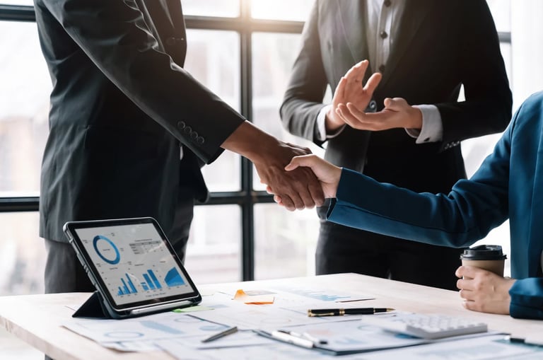 Business professionals shaking hands over a desk with a tablet displaying charts and financial documents
