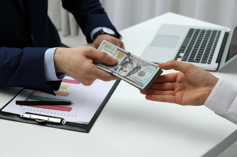 Person in business suit handing US dollar bills to another person at a desk with laptop and clipboard